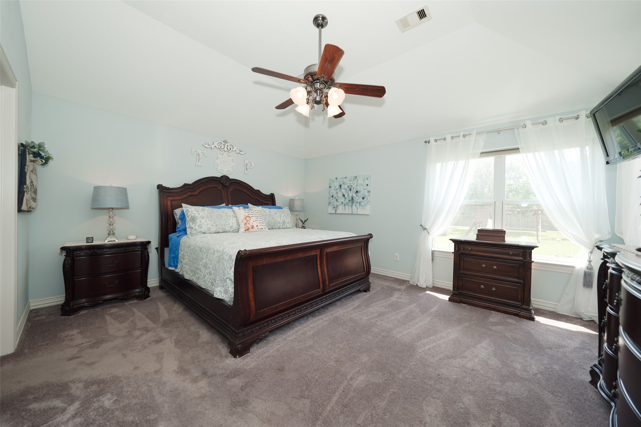 2203 Red Bird Lane Pattison, TX 77423 - Photo 19 of 46 This spacious bedroom features a large window with sheer curtains, allowing natural light to fill the room. It includes a ceiling fan, elegant dark wood furniture, and plush carpeting, creating a cozy and inviting atmosphere.