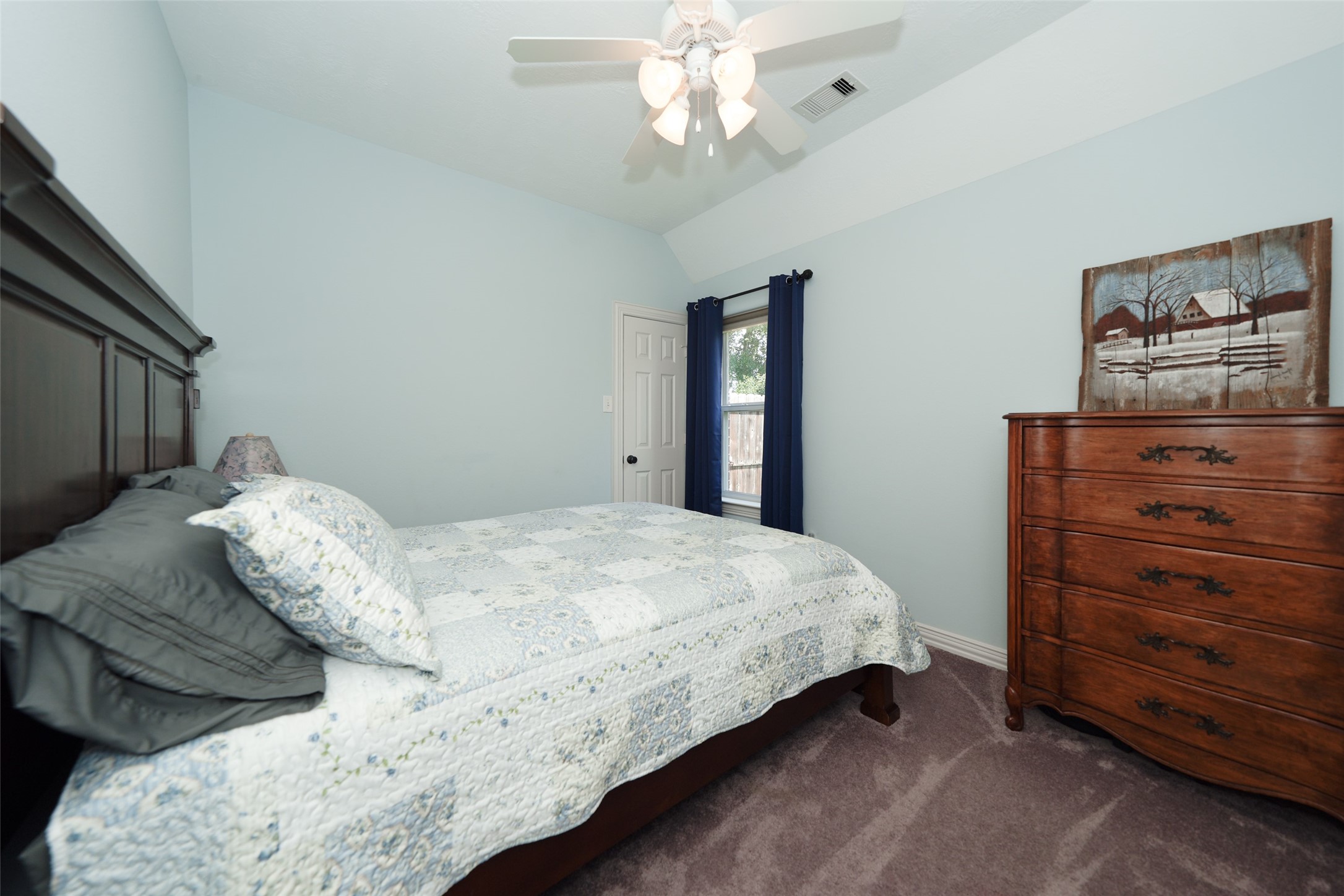2203 Red Bird Lane Pattison, TX 77423 - Photo 28 of 46 This cozy bedroom features a vaulted ceiling with a ceiling fan, soft blue walls, and plush carpeting. It includes a large wooden bed, matching dresser, and a window with dark curtains, offering natural light and a peaceful ambiance.