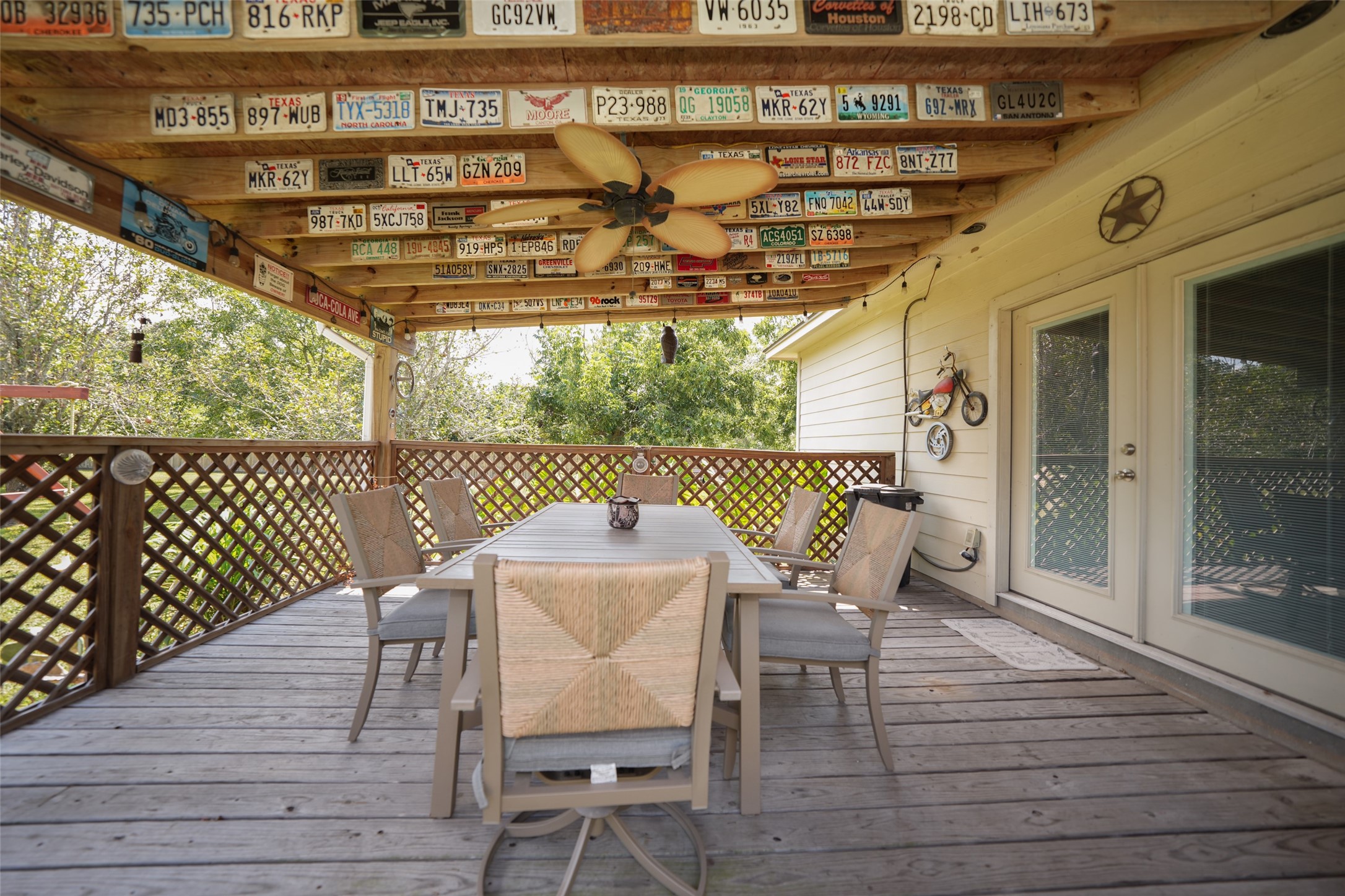 2203 Red Bird Lane Pattison, TX 77423 - Photo 38 of 46 This photo showcases a charming covered patio with a rustic wooden deck. The ceiling is adorned with vintage license plates, adding character. There's a spacious dining area with comfortable seating, perfect for outdoor gatherings. Lattice railing provides a cozy and inviting atmosphere, with lush greenery visible in the background.