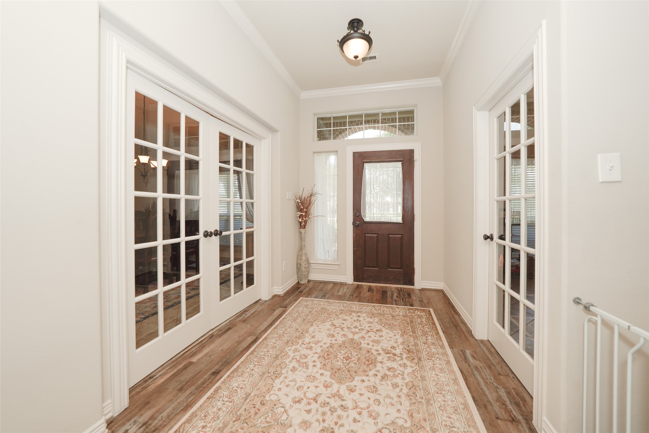 2203 Red Bird Lane Pattison, TX 77423 - Photo 7 of 46 This photo showcases a bright, welcoming entryway with a dark wood front door, sidelights, and a transom window. The space features elegant French doors on both sides, a stylish light fixture, and a patterned area rug on the hardwood floor, creating a warm and inviting atmosphere.