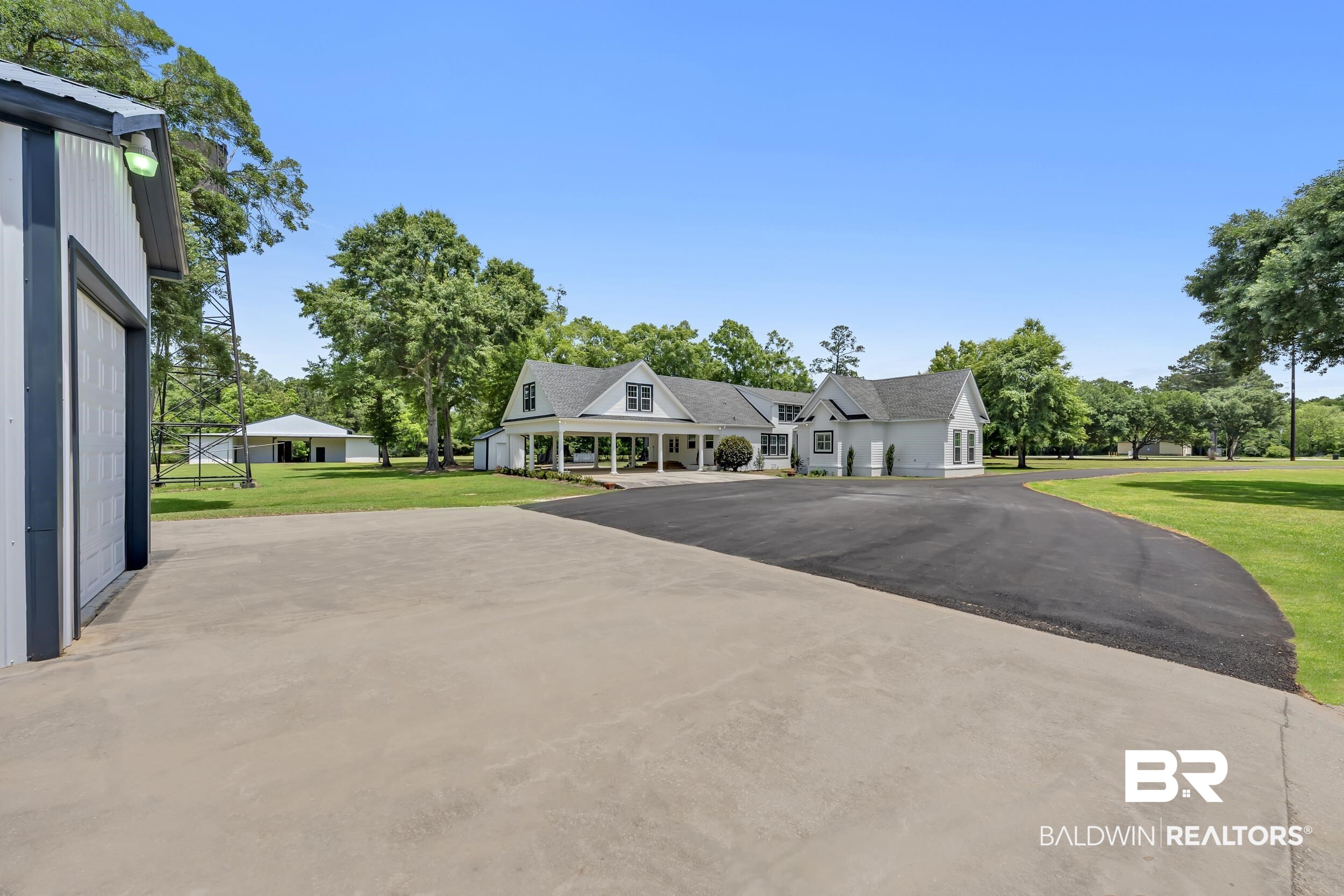 43199 Pine Grove Road Bay Minette, AL 36507 - Photo 5 of 71 White exterior residence with a covered porch and portico, featuring a detached garage, asphalt driveway, and concrete parking pad, all set against a backdrop of mature trees and expansive lawn