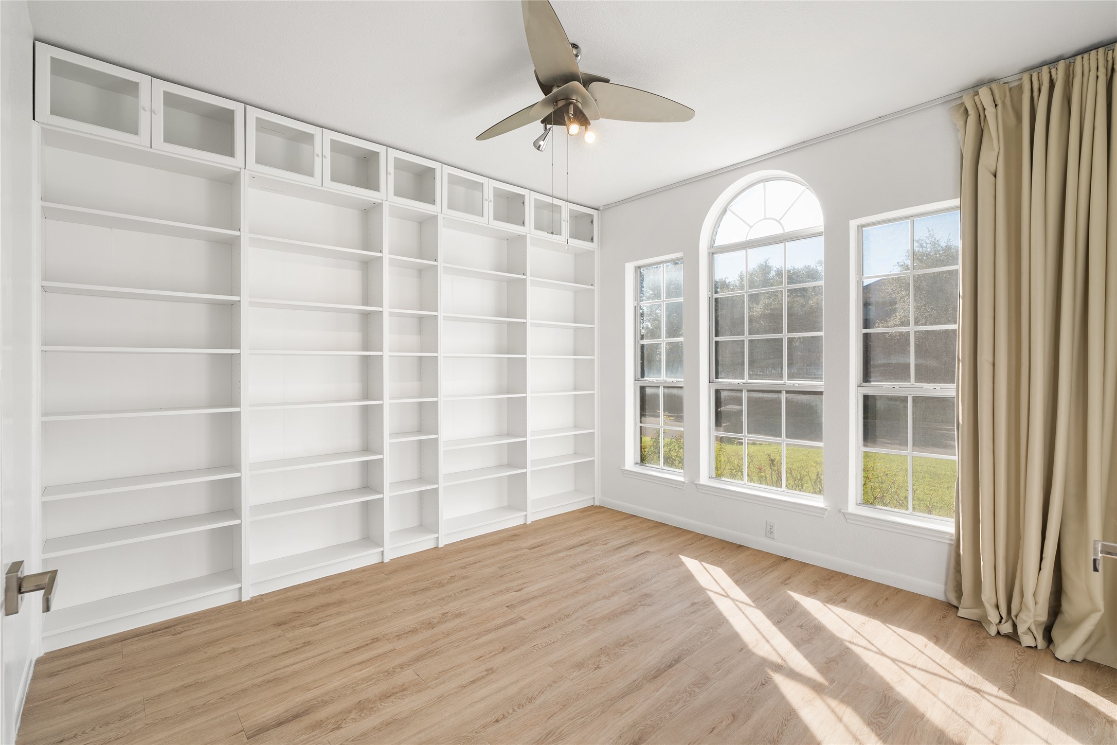 206 River Down Road Georgetown, TX 78628 - Photo 8 of 39 Extensive built-in shelving with glass-front upper cabinets, wood-finish flooring, a ceiling fan, and multiple windows including an arched top window