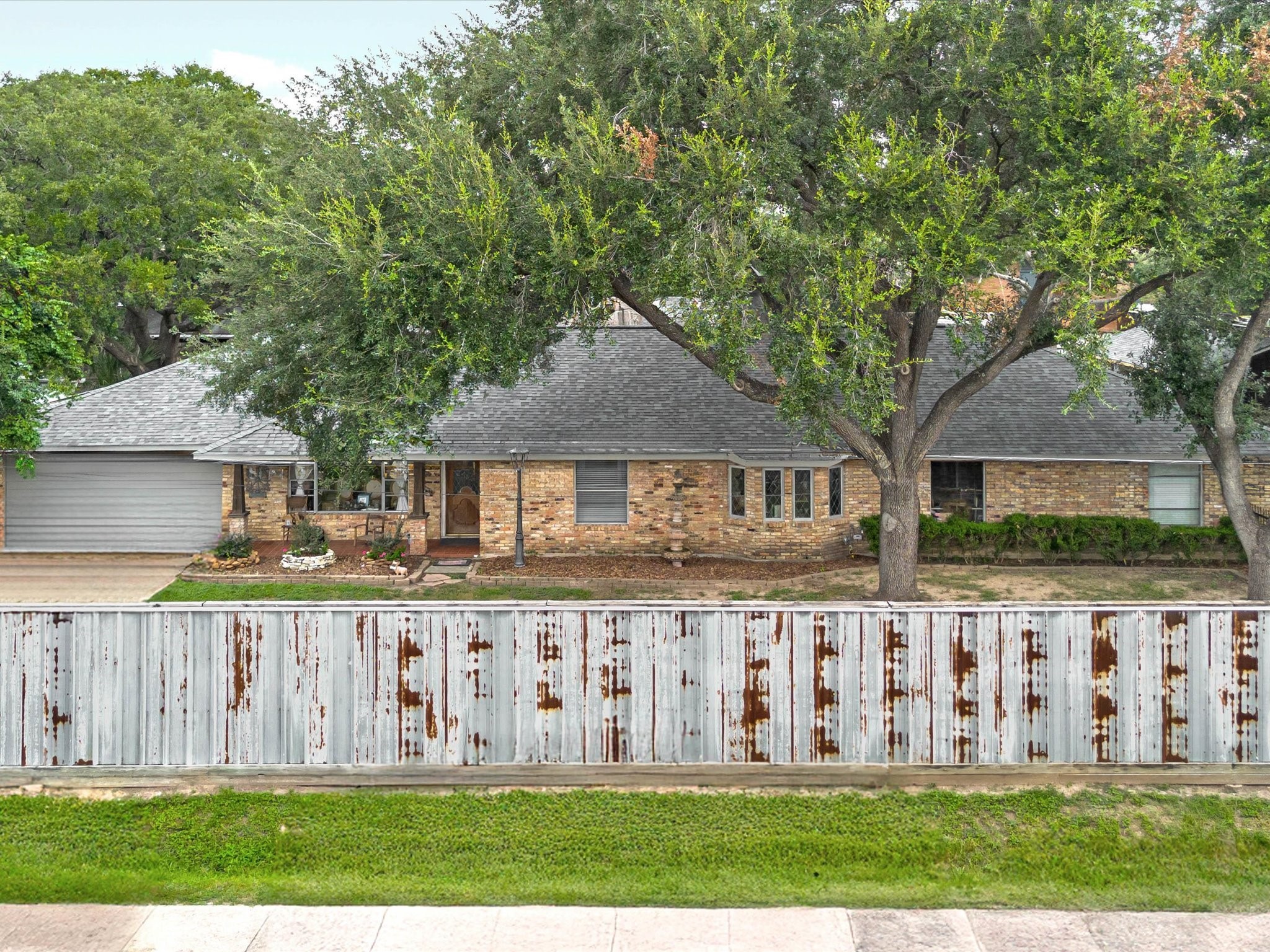 This charming single-story traditional home features a classic brick exterior framed by mature shade trees. Large front-facing windows invite natural light inside, while a rustic metal privacy fence adds character and seclusion from the street.