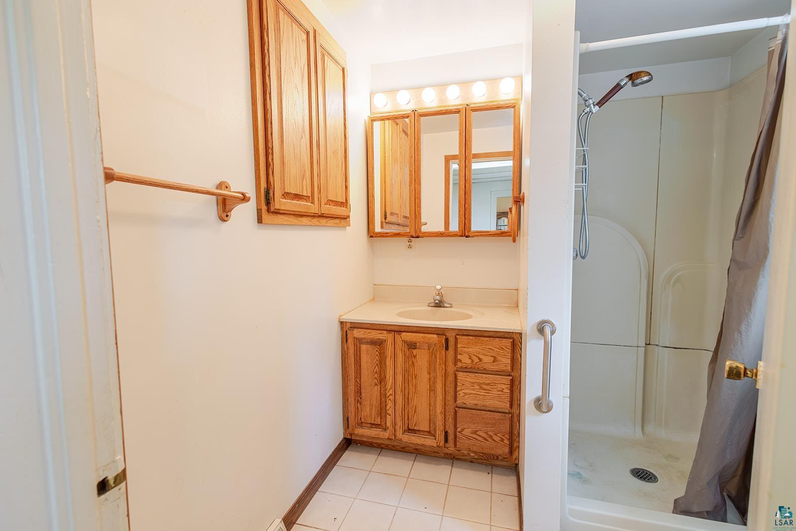 438 9th Street Cloquet, MN 55720 - Photo 15 of 27 Bathroom featuring a wood-finish vanity with an integrated sink and light-toned countertop, a mirrored medicine cabinet with overhead lighting, a standing shower, and white tile flooring