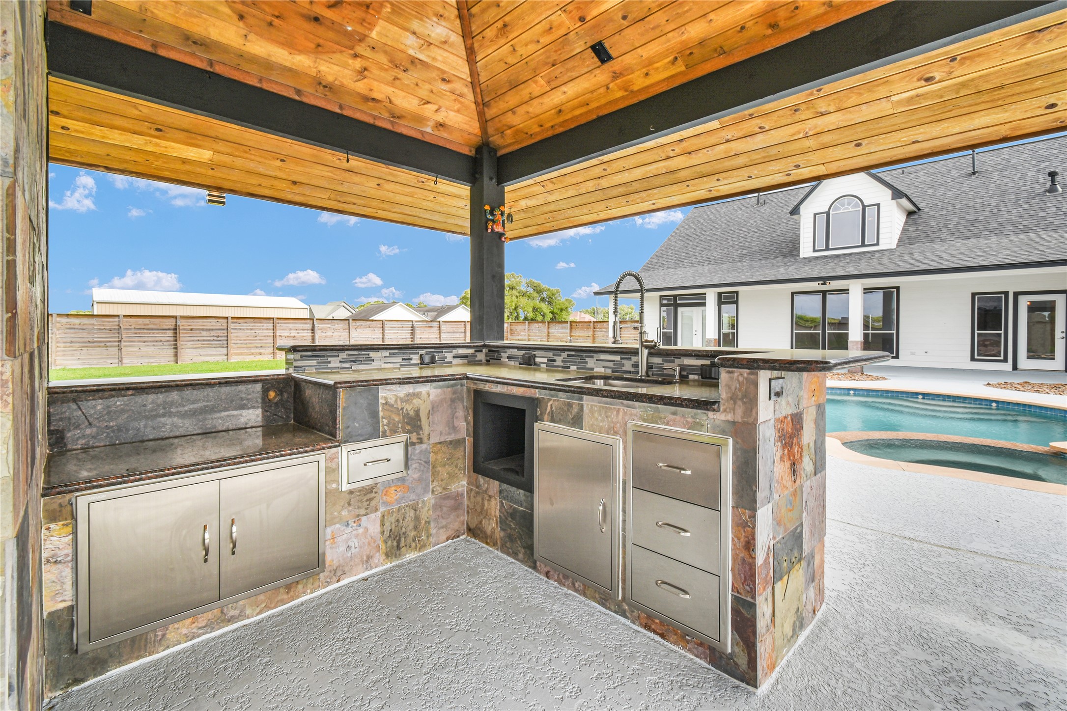 863 Bunkhouse Trail Angleton, TX 77515 - Photo 18 of 22 This photo showcases a stylish outdoor kitchen with a slate tile finish, stainless steel appliances, and a sink, all under a wood-paneled ceiling. Adjacent is a sparkling pool,and a spacious patio visible in the background. Perfect for entertaining and outdoor living.